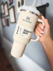 Beige Stanley mug with handle held by a hand against a blurred indoor background